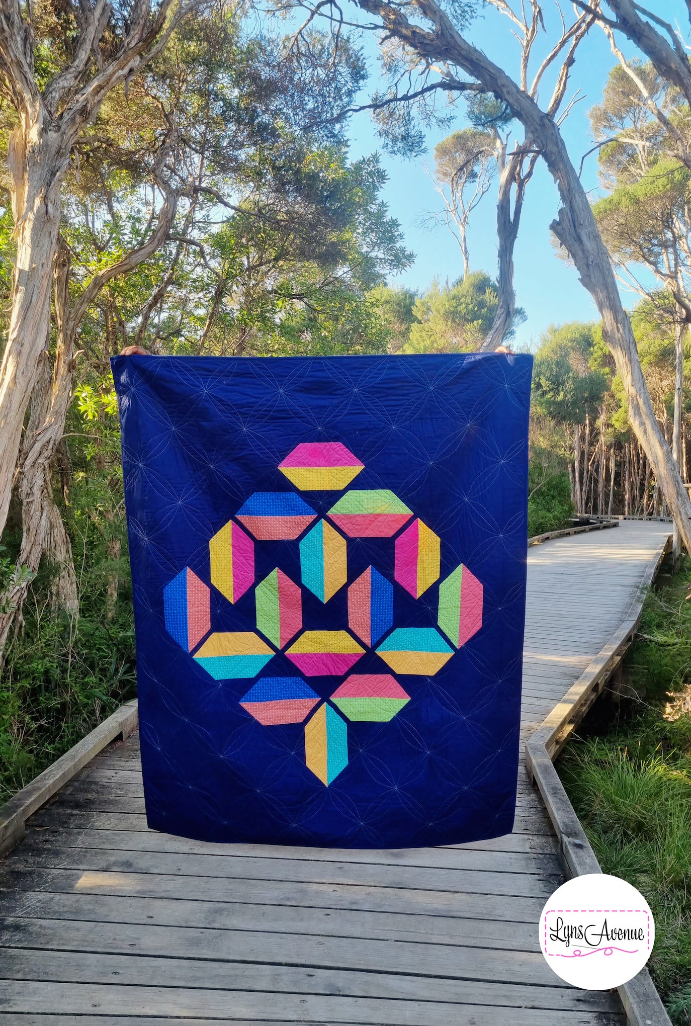 Flourishing quilt pattern in scrappy colours pink, peach, yellow, blue, green and lime on a navy blue background with trees in the background